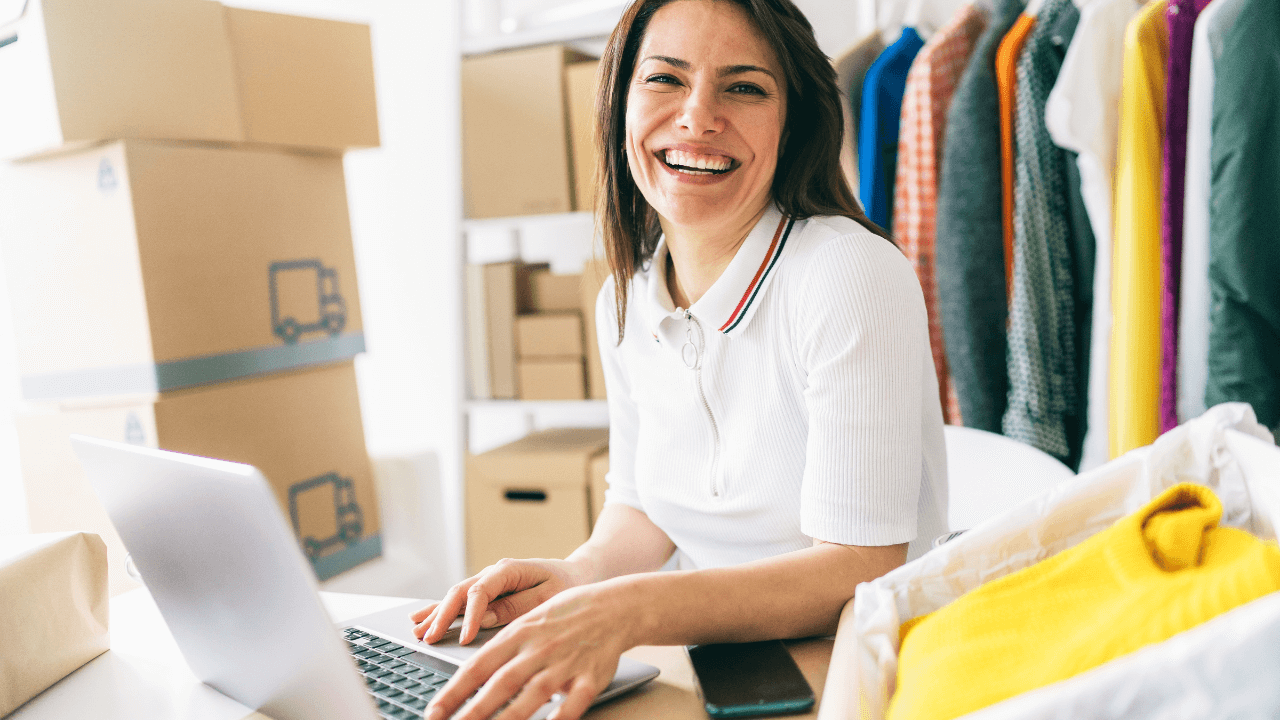 Smiling small business owner running an online shop from her laptop, surrounded by shipping boxes and clothing stock