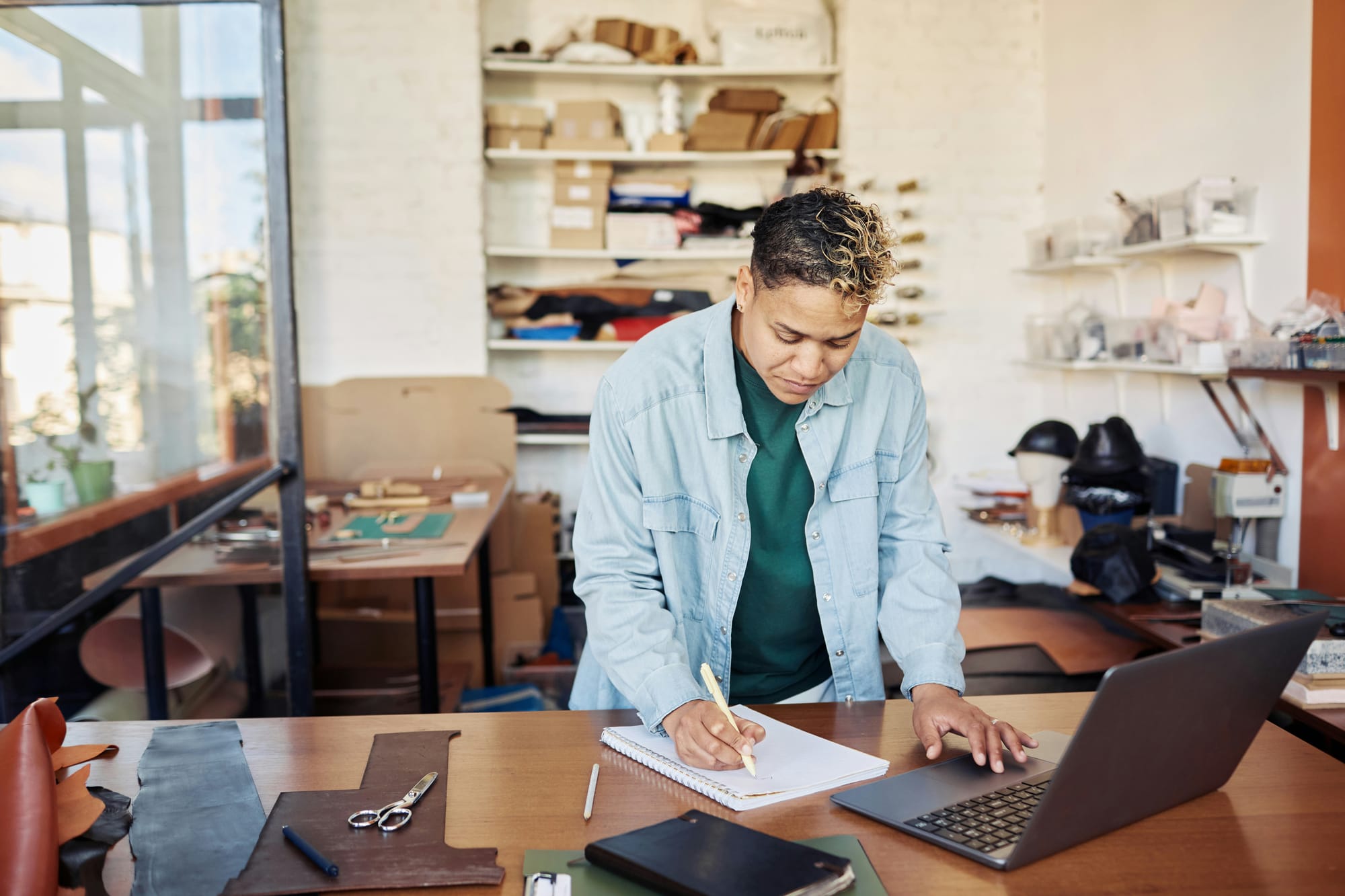 Small business owner writing in a notebook while checking her laptop in a leather goods workshop