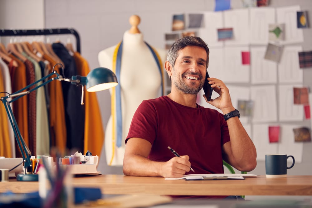 Smiling small business owner on the phone at his desk, surrounded by fabric swatches and a tailor's mannequin